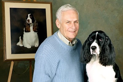 Charles L. "Bud" Kramer posing in a blue sweater with his English Springer Spaniel in front of a painting of a dog